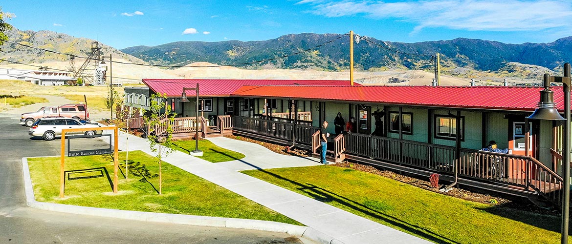 The exterior of a long, one-story building with a red roof and mountains and blue sky in the background